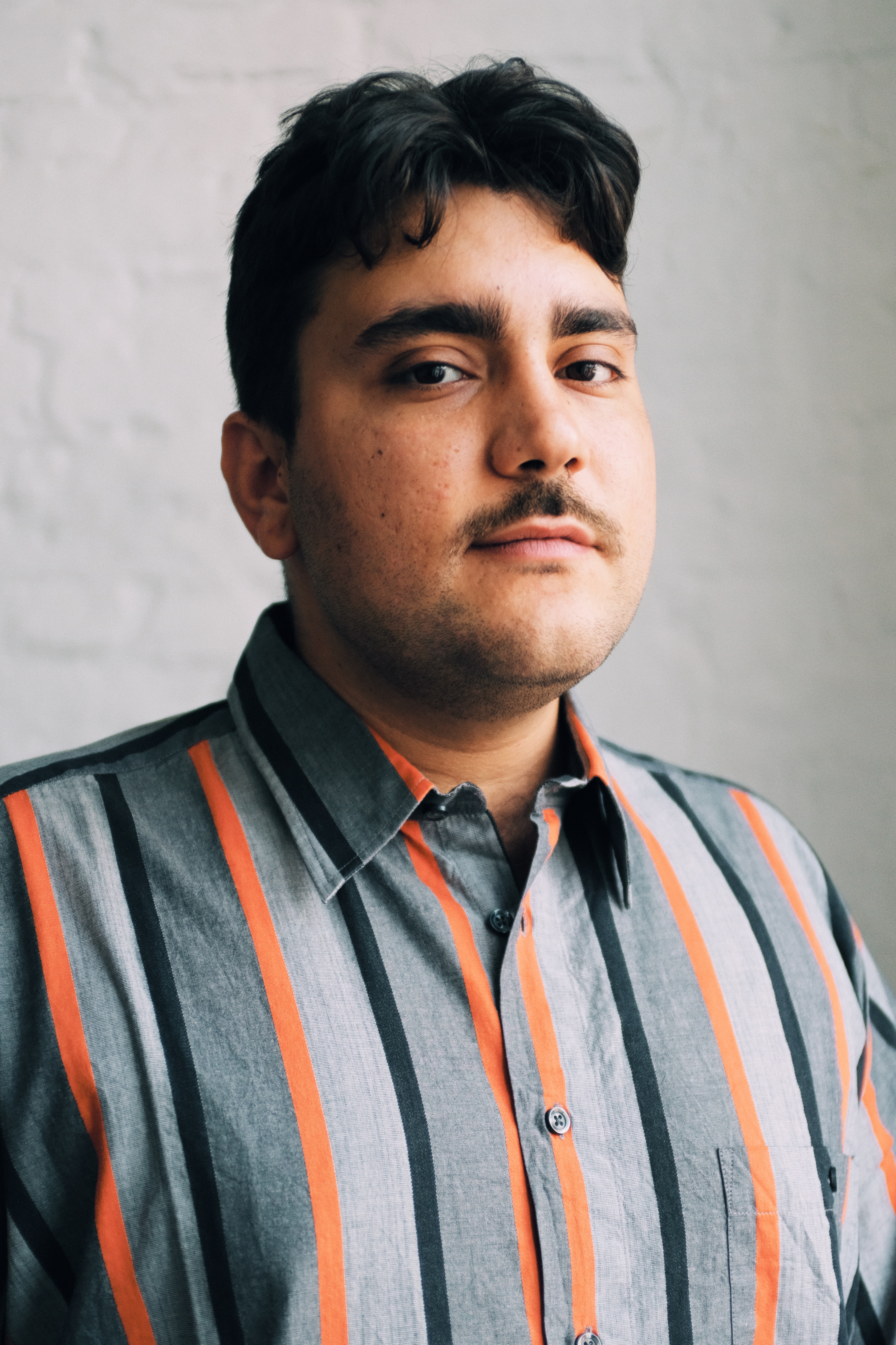 Portrait of a man with a grey and orange strippy shirt, thin black moustache and thick dark hair looks at the camera