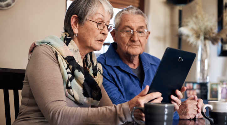 Two older people sit at a table looking at a tablet